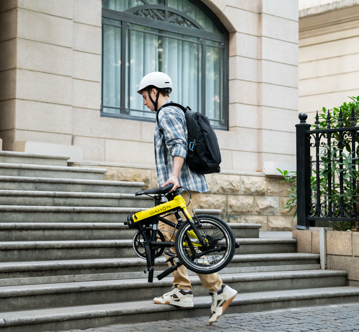 A man in casual attire carries a folded yellow DAHON bike up stone steps, showcasing its portability and urban versatility.