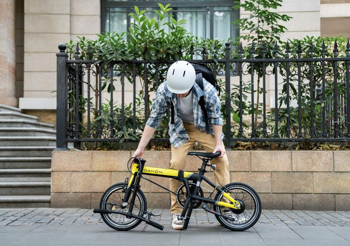 A person wearing a helmet and casual clothing is adjusting or inspecting a yellow folding bicycle on a sidewalk in front of a building with a black iron fence.