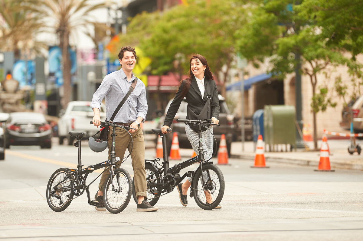 A man and a woman walk with their DAHON folding bikes on a city street, showcasing urban commuting style.