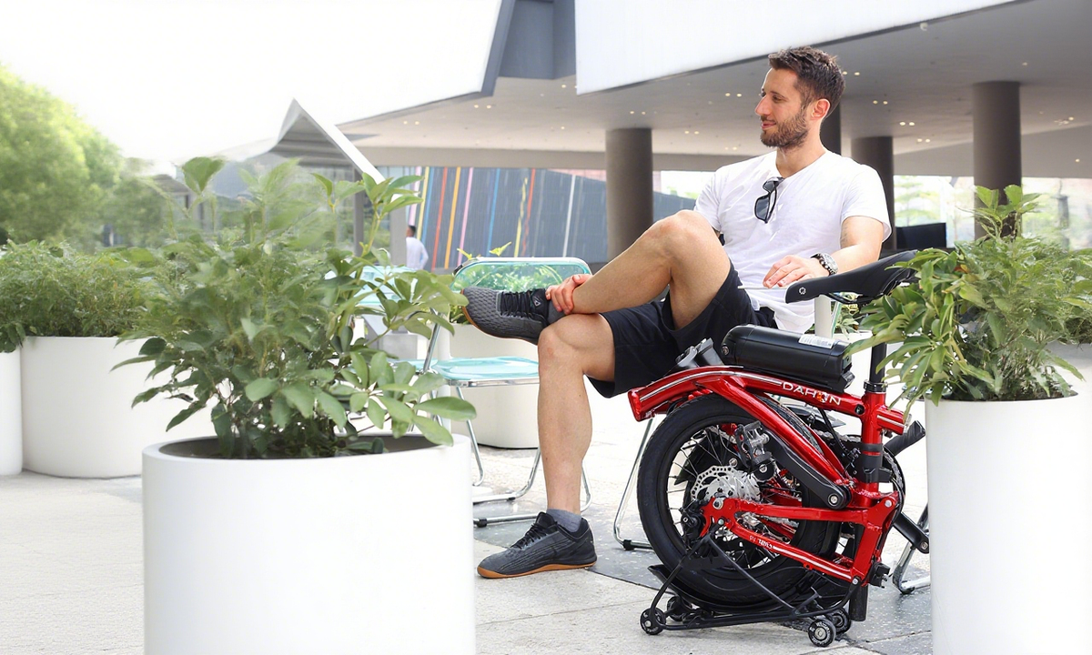 A man relaxes on a bench with a red DAHON folding bike beside him in an outdoor urban setting.