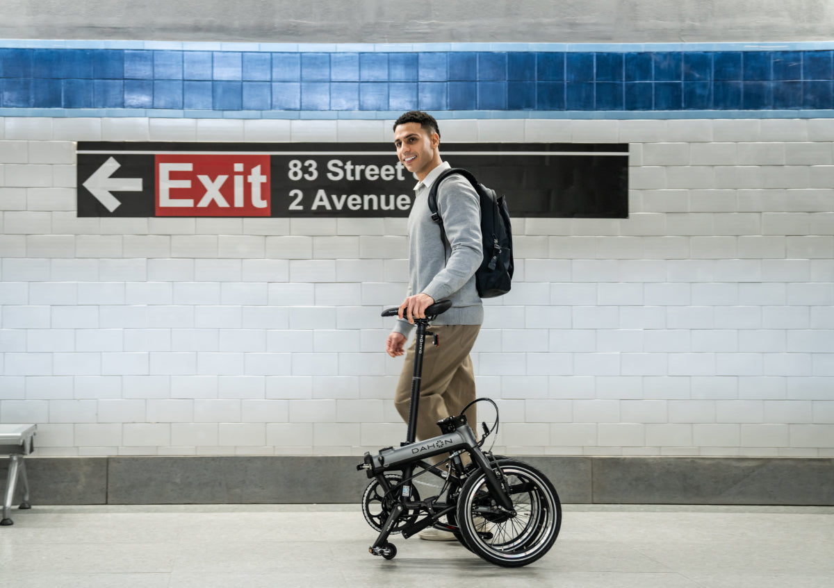 Man with folded dark grey Dahon K-Feather e-bike in subway station, showcasing urban commuting convenience.