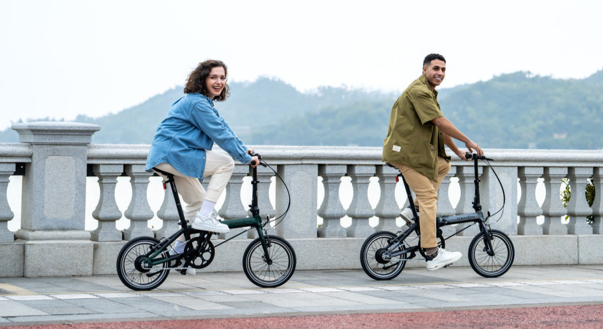 Couple riding dark green and black DAHON K-Feather folding e-bikes on a scenic path, highlighting portability and urban lifestyle.